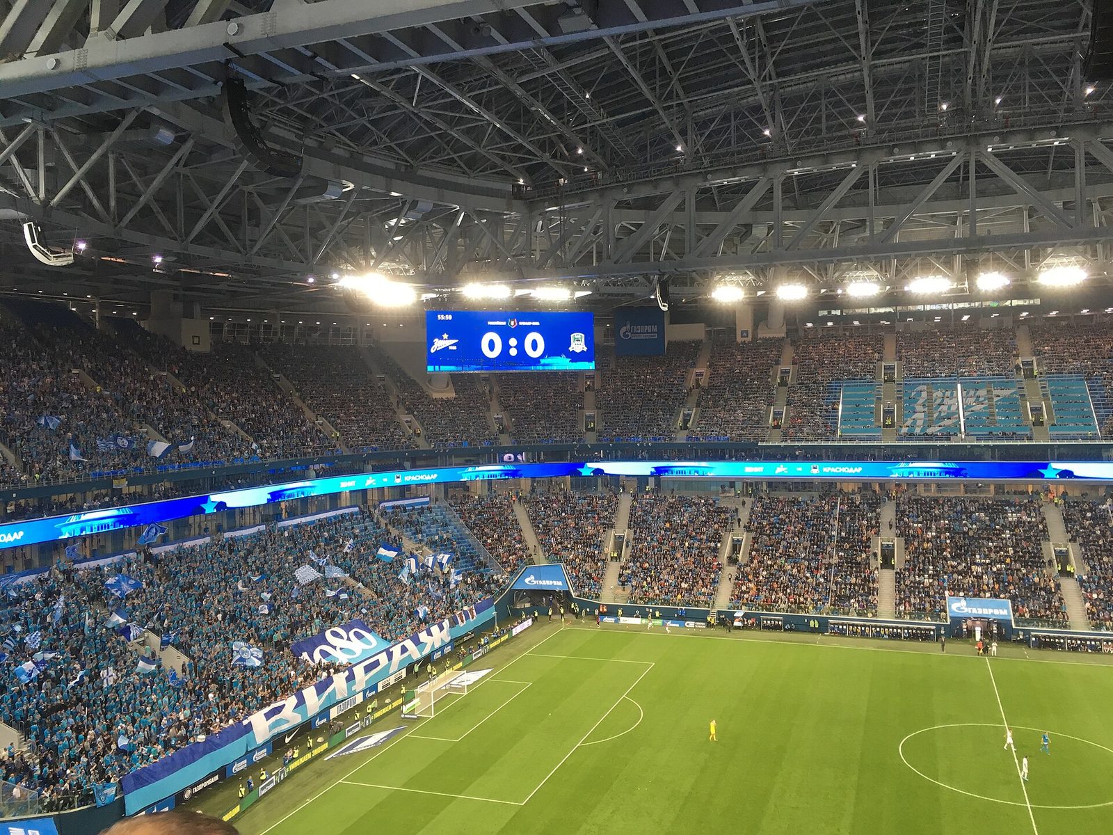 Заполненный футбольный стадион под крышей, на табло счёт 0:0, болельщики с флагами на трибуне